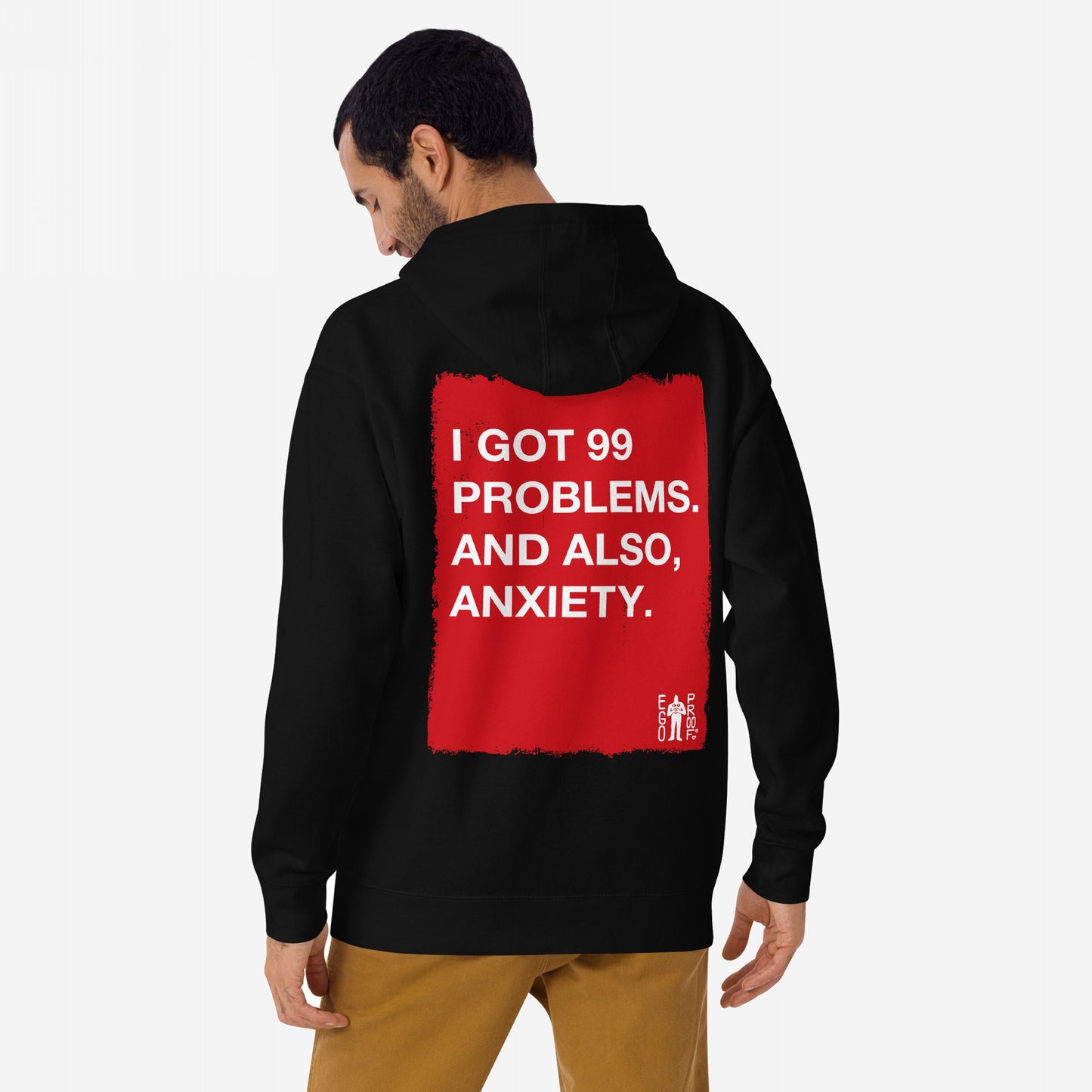 Person wearing the 99 Problems Hoodie EU, featuring bold white text on a red rectangle: I GOT 99 PROBLEMS. AND ALSO, ANXIETY. Small EGO PROOF logo in the corner. Light brown pants complement the look.