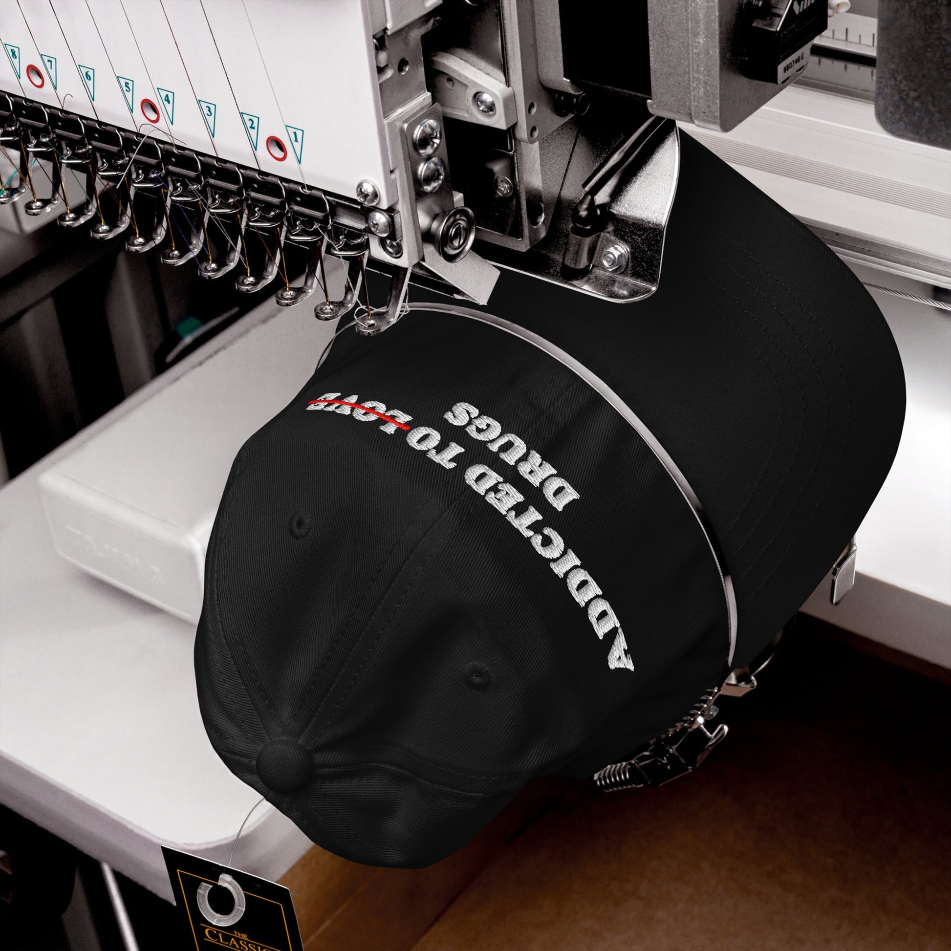 A machine stitches a black Egoproof snapback, the Addicted To Love / Drugs Hat, featuring bold white and red embroidered text—a striking mental health statement cap in the making.