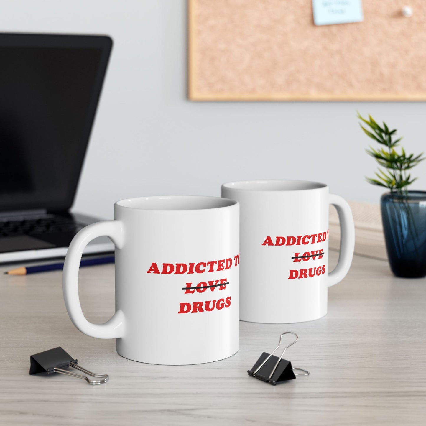 Two white ceramic mugs featuring the slogan Addicted To ~Love~ Drugs in red letters on a wood desk, surrounded by office items, embodying dark humor and mental health awareness.