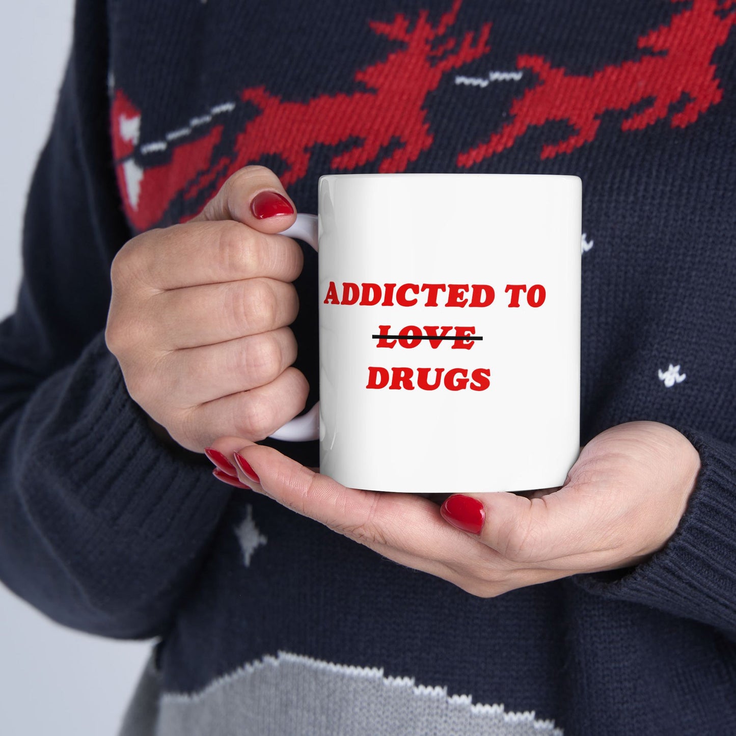 Hands with red nails hold the Addicted To Love / Drugs Mug, featuring bold red text humorously contrasting love and medication. The mug complements a dark sweater with a red reindeer pattern.