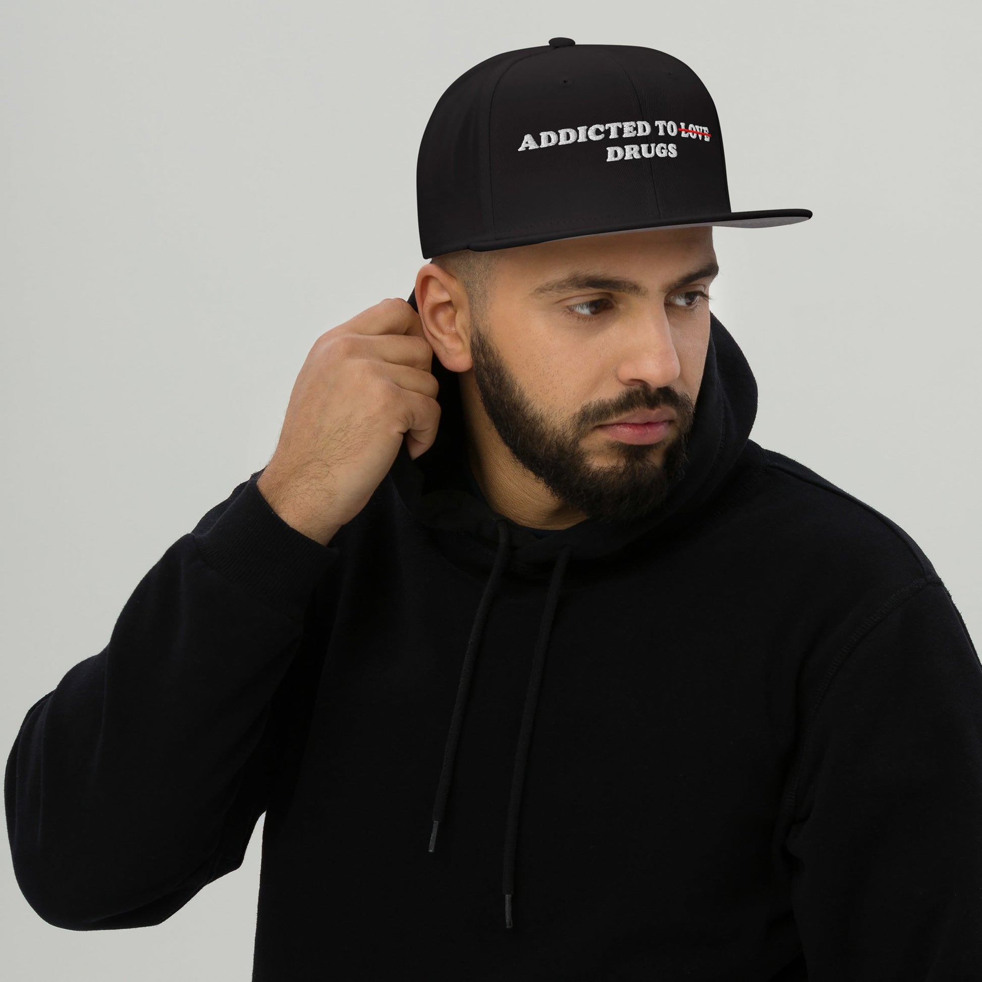 A man in a black hoodie and an Egoproof Addicted To Love / Drugs Hat looks to the side, touching his neck. The embroidered snapback makes a bold mental health statement against a plain light background.