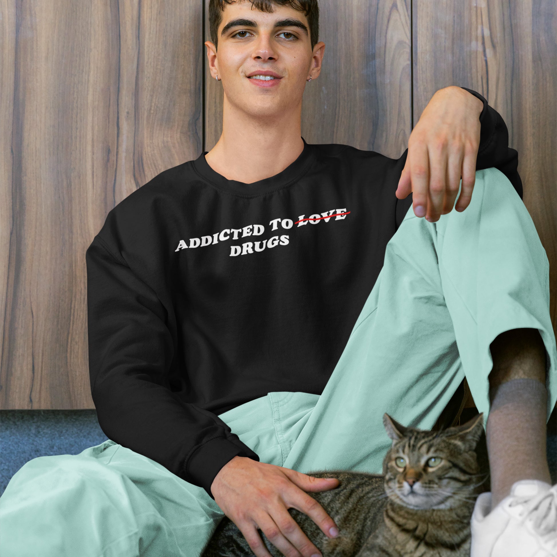 Young man wearing Addicted To Love / Drugs Hoodie & Sweatshirt in black, sitting relaxed with a cat on his lap against wood panel background