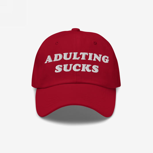 Red baseball-style cap with ADULTING SUCKS embroidered in white on front, representing Egoproof's relatable, bold apparel. Ideal for expressing millennial and Gen Z frustrations with style.