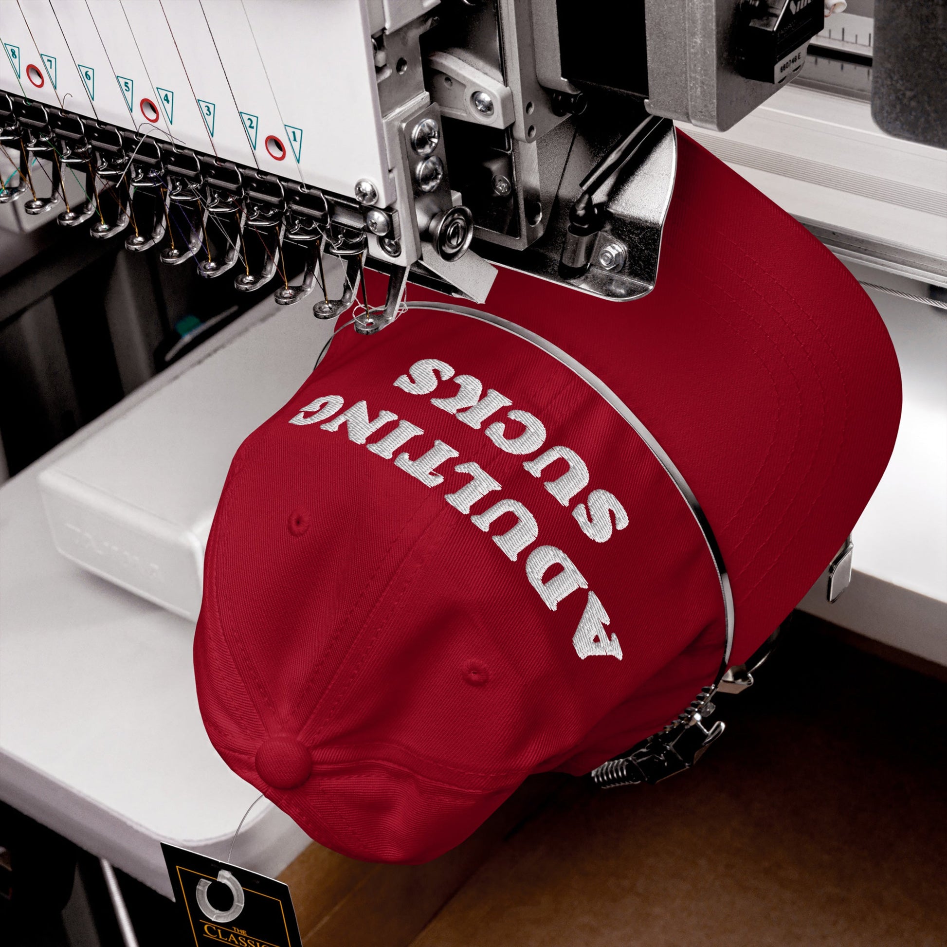 A red Adulting Sucks hat is clamped in a multi-needle embroidery machine, stitching the slogan in white block letters across the front, reflecting Egoproof's honest, relatable message.