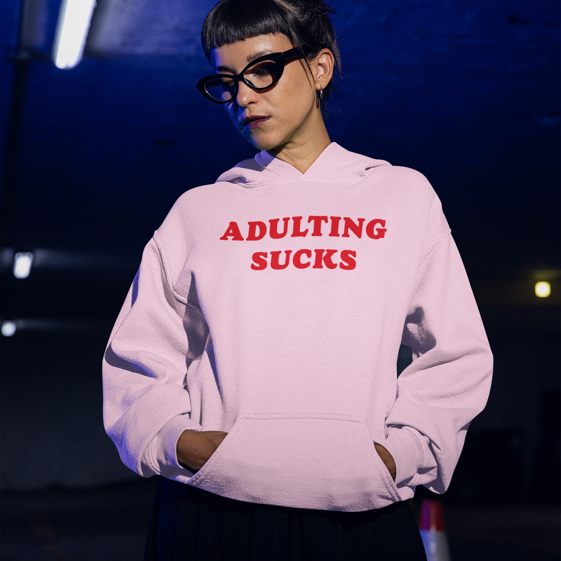 Adulting Sucks Hoodie worn by a person in glasses, featuring bold red text on a light pink comfy midweight hoodie.