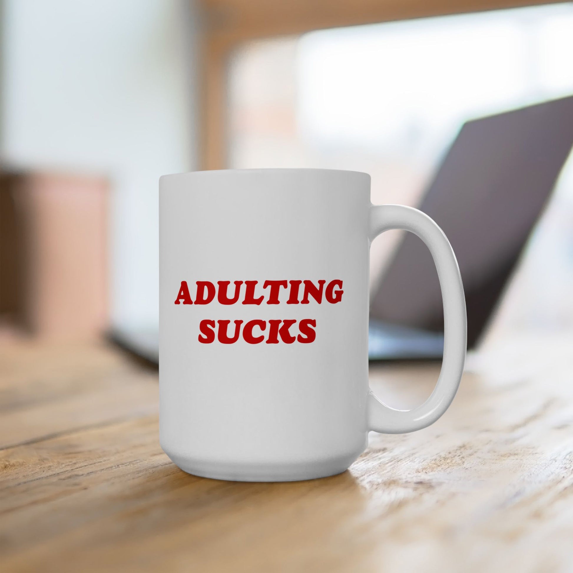 Adulting Sucks Mug on a wooden tabletop, showcasing bold red lettering. Ideal for office rebels, this ceramic mug humorously captures life's daily grind, perfect for those embracing life's chaos.