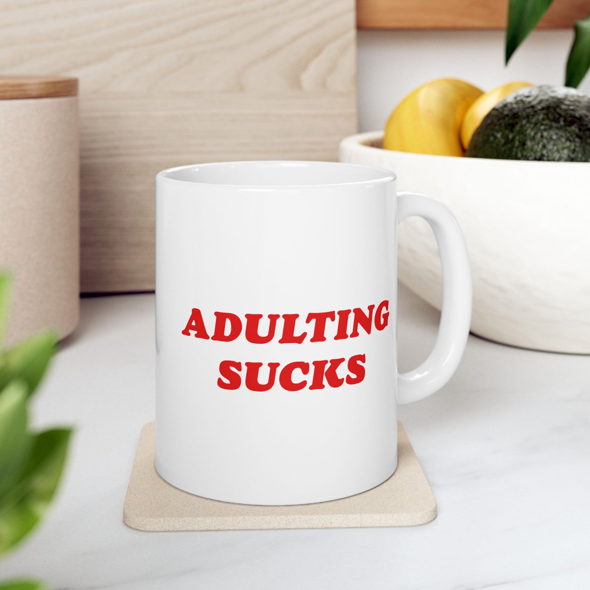 Adulting Sucks Mug on a coaster, showcasing bold red lettering. Ceramic design fits artful rebellion, ideal for those facing daily challenges. Accompanied by fruit and plant, embodying Egoproof’s strong, relatable expressions.