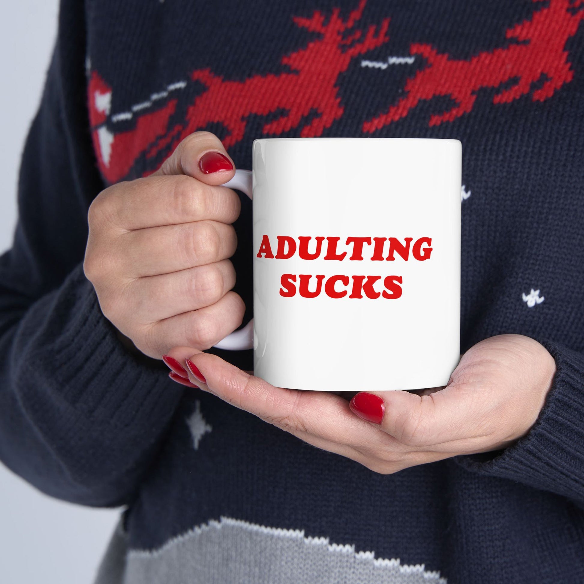 A person holds the Adulting Sucks Mug, featuring bold red text, cradled in hands with red nail polish. This white ceramic mug is perfect for expressing daily frustrations.