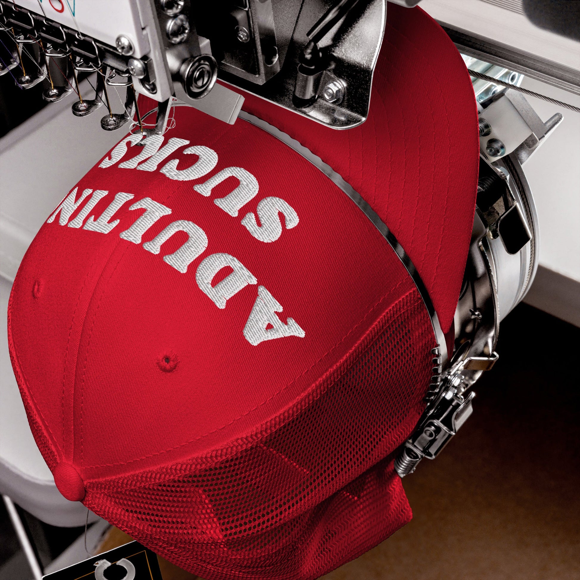 The image shows an Adulting Sucks Hat in red mesh-back style, clamped in an embroidery machine, with white thread stitching the phrase ADULTING SUCKS on the front panel.