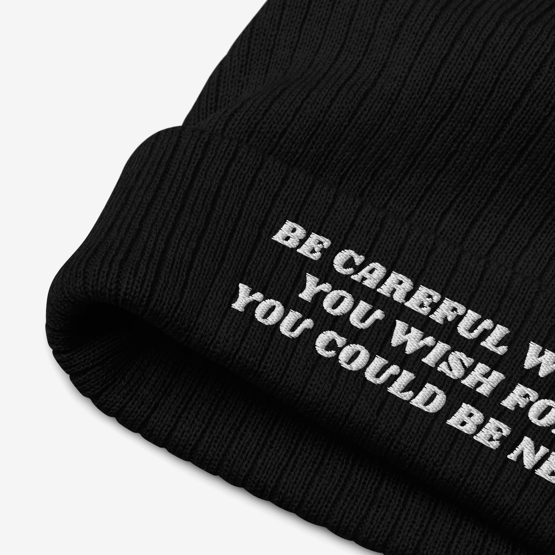 Black rib-knit winter beanie with folded cuff style, showcasing white embroidered text: BE CAREFUL WHAT YOU WISH FOR – YOU COULD BE NEXT. Known as the Be Careful Hat, it reflects bold, progressive statements.
