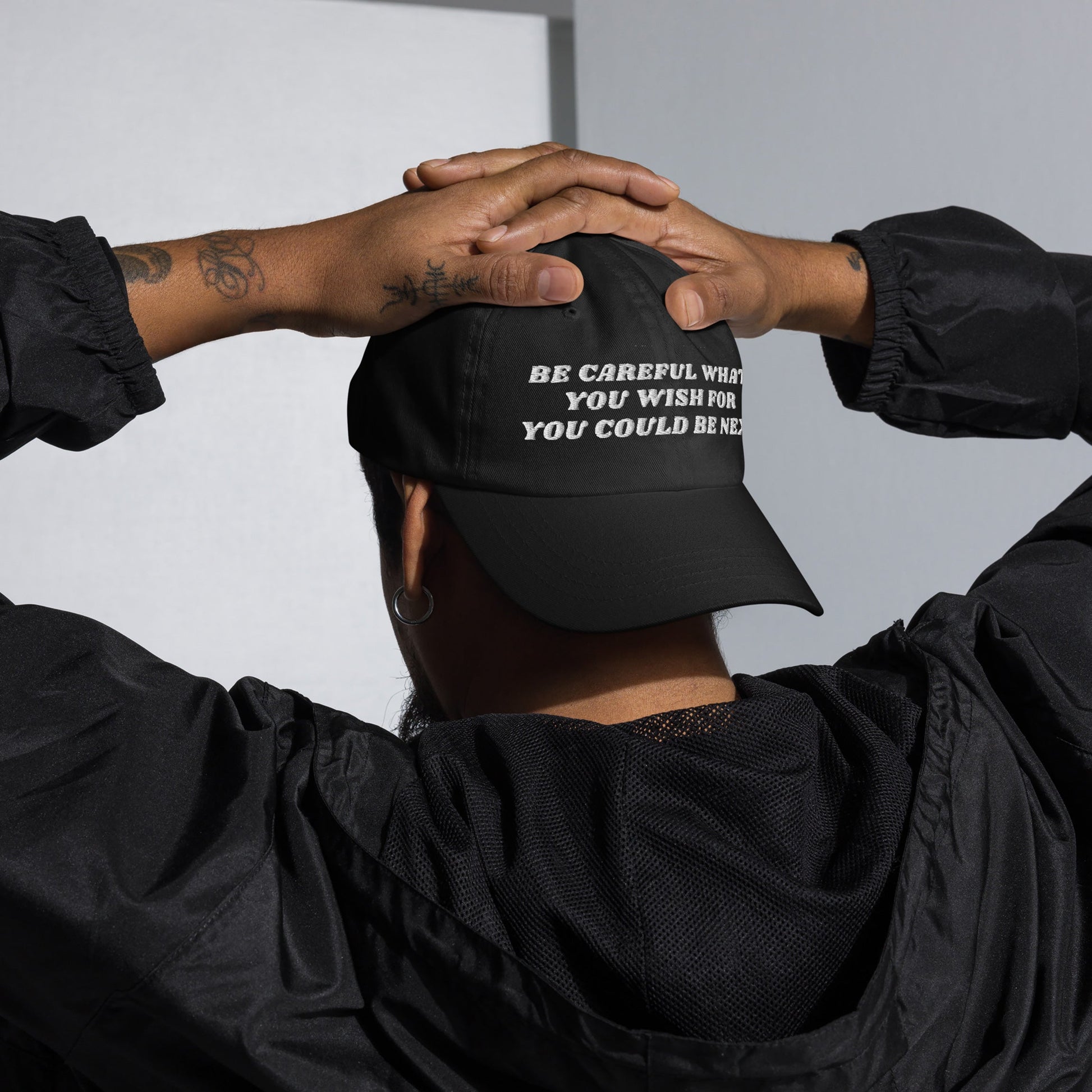 Person wearing the Be Careful Hat, a black cap with bold white embroidery. Their hands reveal forearm tattoos, embodying a defiant statement. Ideal for anti-fascists and progressives.