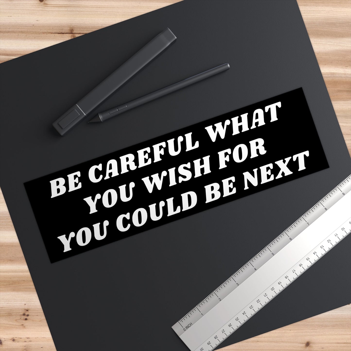 Top-down view of a wood-grain desk featuring the Be Careful Sticker & Magnet with bold white text warning, “BE CAREFUL WHAT YOU WISH FOR YOU COULD BE NEXT,” alongside a digital pen and metal ruler.