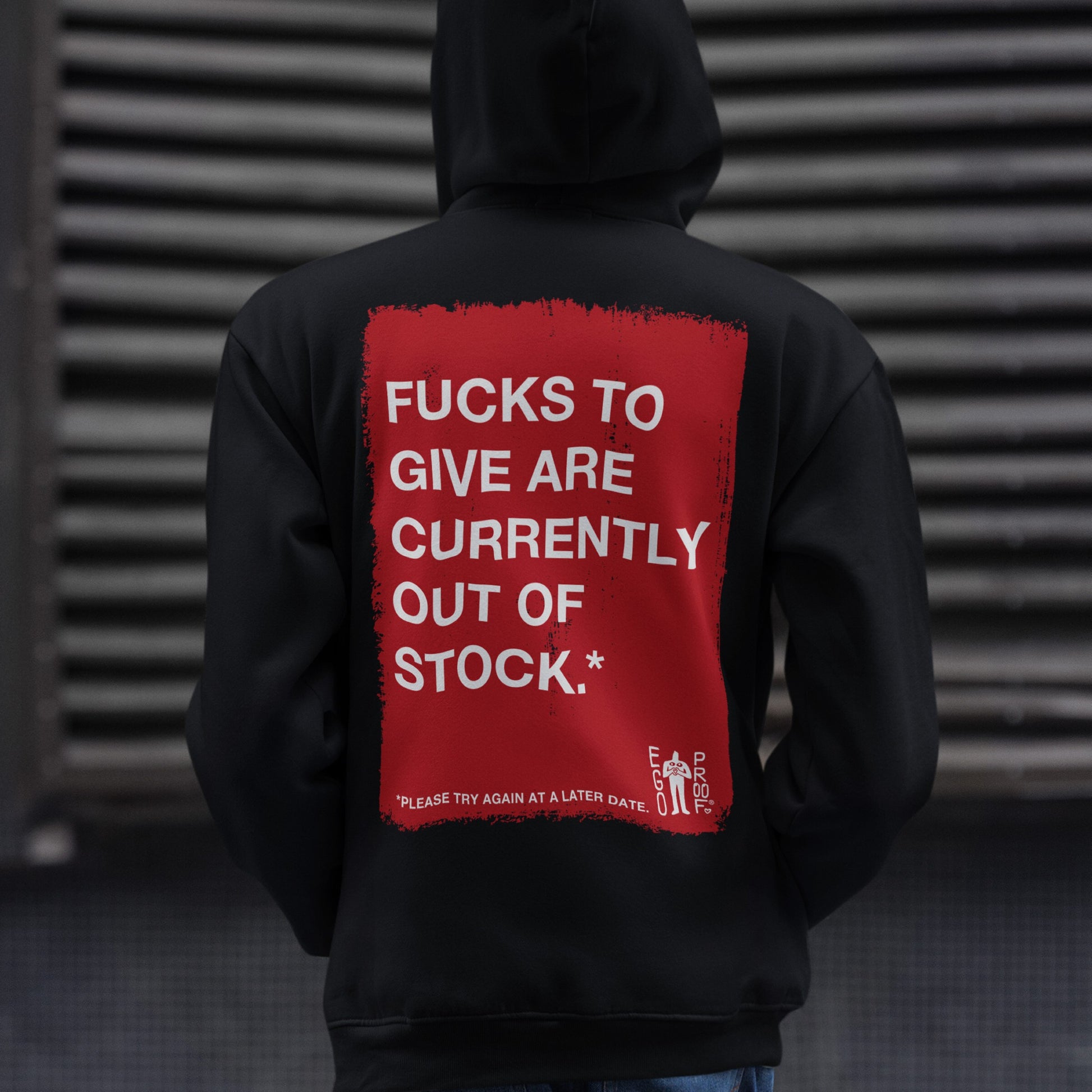 Person wearing the Fucks To Give Hoodie, featuring a distressed red rectangle with bold white text on the back, standing against an industrial background. The hoodie embodies Egoproof's theme of unapologetic self-expression.