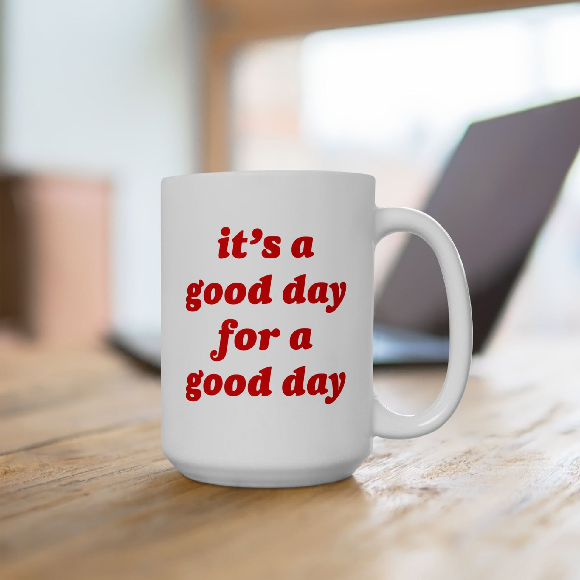 White ceramic Good Day Mug on a wooden desk, featuring bold red text: “it’s a good day for a good day.” Background hints at a casual work setting with a blurred laptop.
