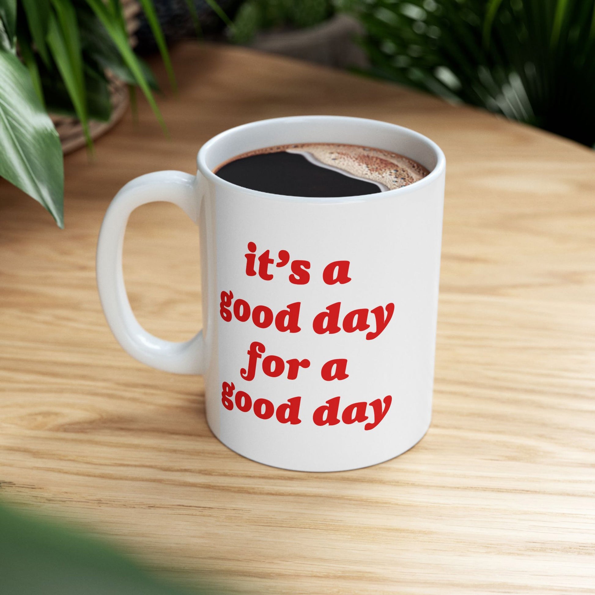 Good Day Mug on a light-wood tabletop, filled with black coffee. Bold red text reads, “it’s a good day for a good day.” Green houseplant leaves visible in the background.