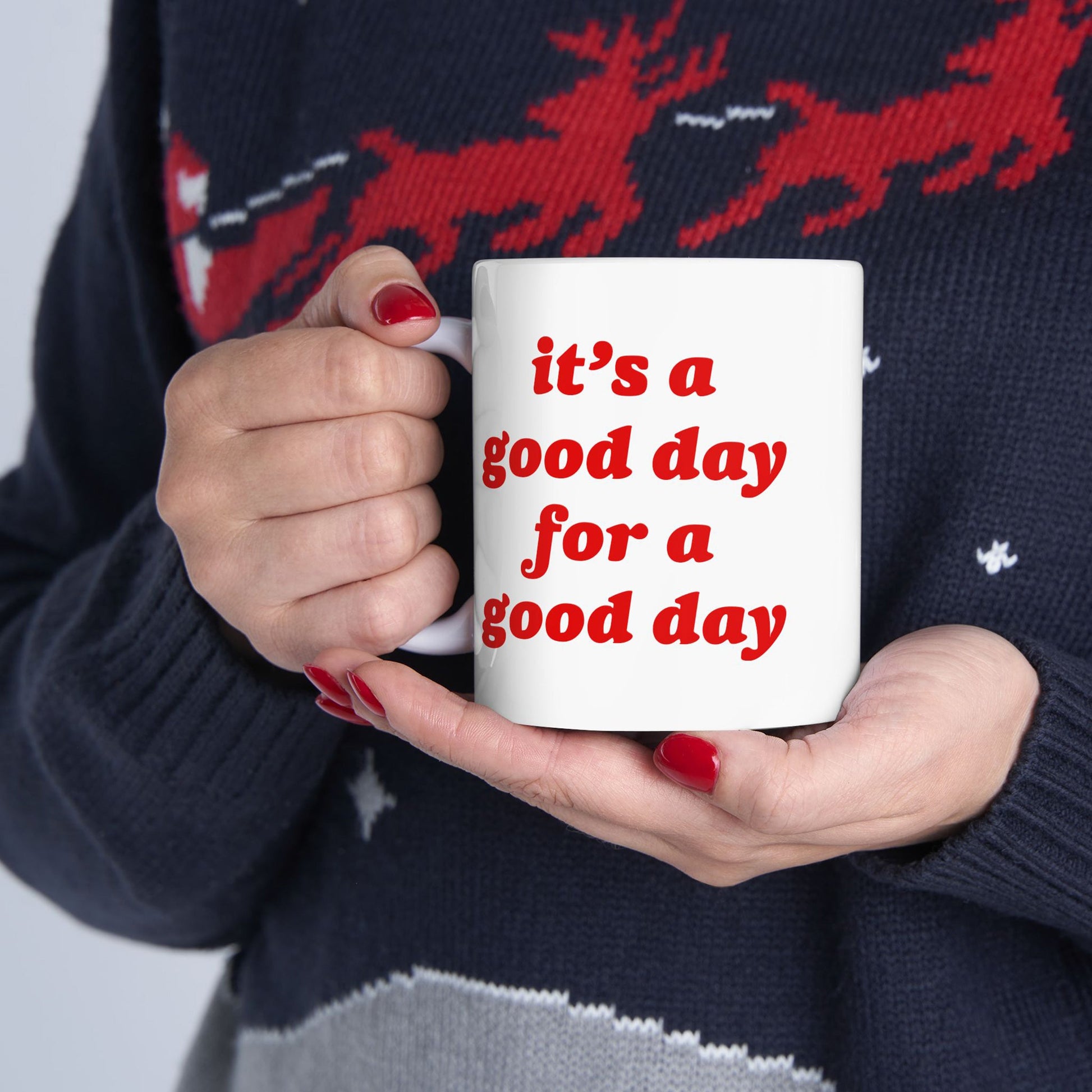 A person cradles the Good Day Mug, adorned with bold red text, it’s a good day for a good day, complementing their red-painted nails and festive dark knit sweater.