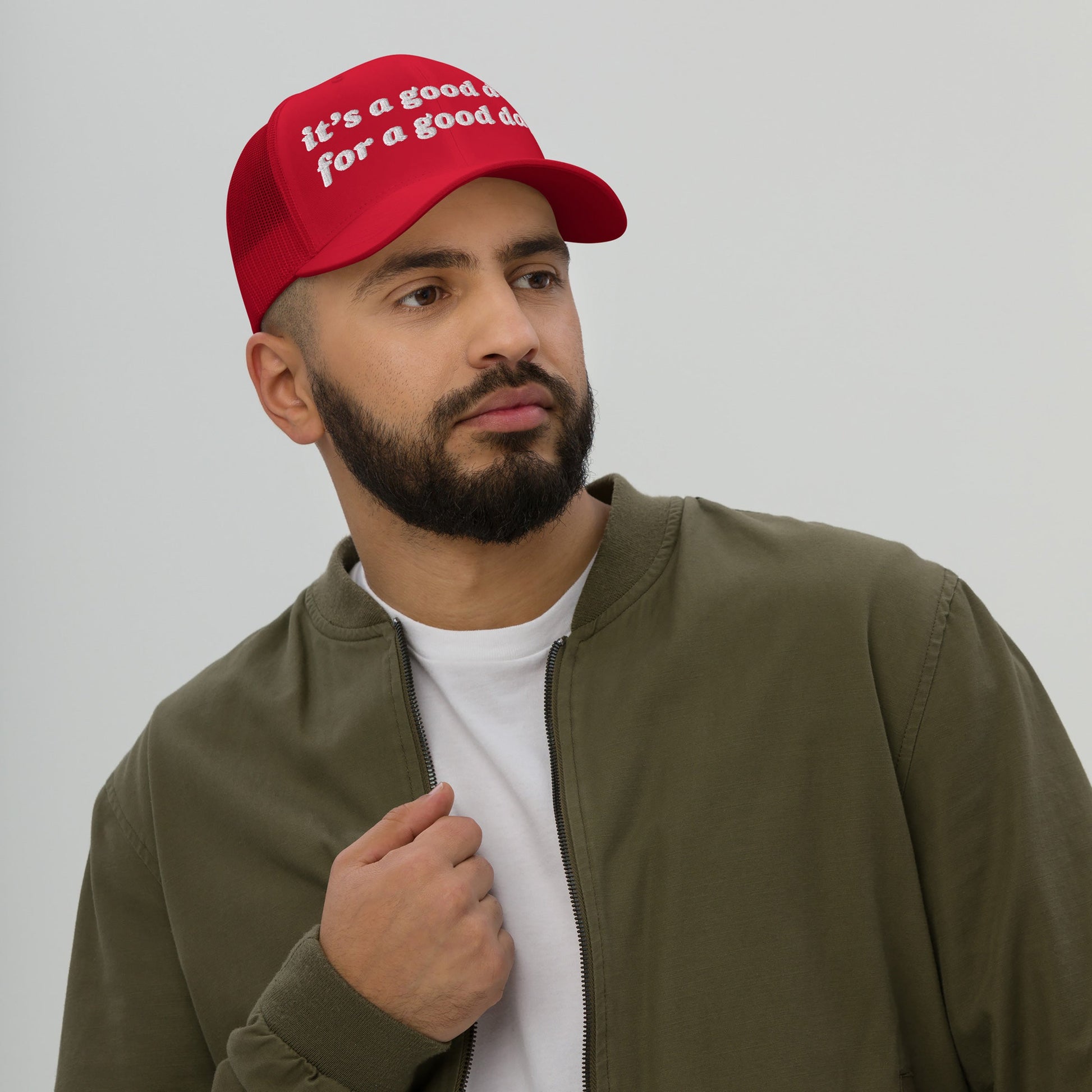 A bearded man wears the Good Day Hat, a bright red mesh-back baseball cap with it's a good day for a good day slogan, styled with an olive bomber jacket and white T-shirt.