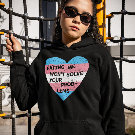 Woman wearing a black No Trans Hate Hoodie with a heart in trans flag colors and the message "Hating Me Won't Solve Your Problems" outdoors.