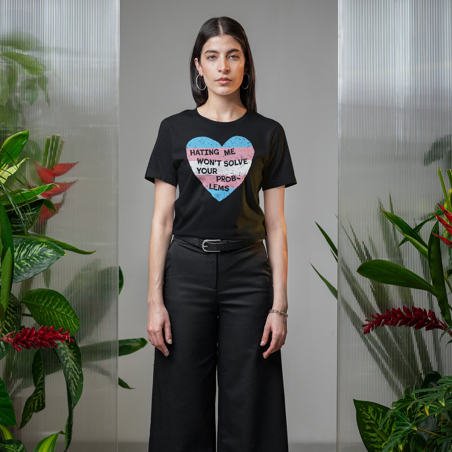 Woman wearing black No Trans Hate T-shirt with trans flag heart and message about hate not solving problems, standing among green plants.