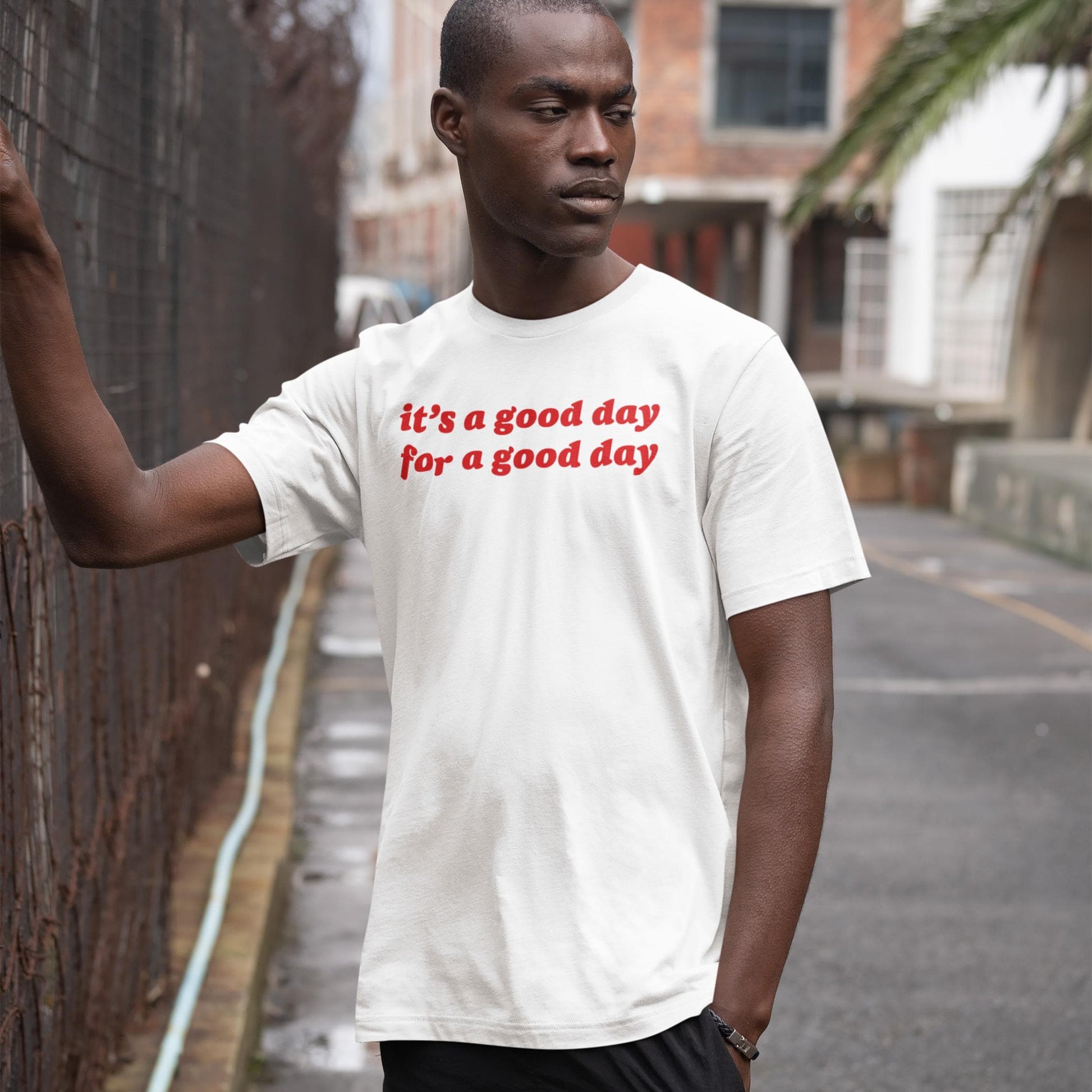 Good Day T-shirt on a young man leaning against a chain-link fence in an urban setting, promoting optimism and mental health awareness with bold red letters.