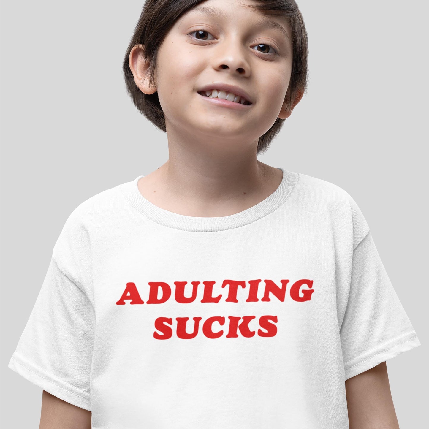 Young boy smiling in a white Adulting Sucks Kids T-shirt EU with bold red lettering, embodying youthful sarcasm and rebellion, symbolizing Egoproof's statement apparel for mental health awareness.