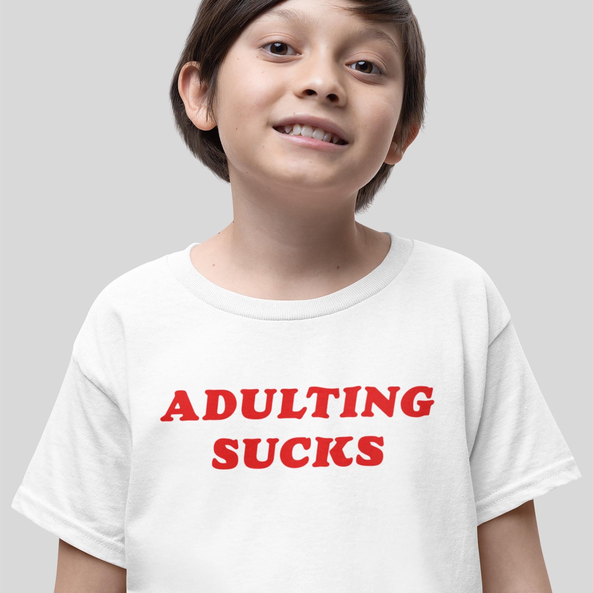 Young boy smiling in a white Adulting Sucks Kids T-shirt EU with bold red lettering, embodying youthful sarcasm and rebellion, symbolizing Egoproof's statement apparel for mental health awareness.