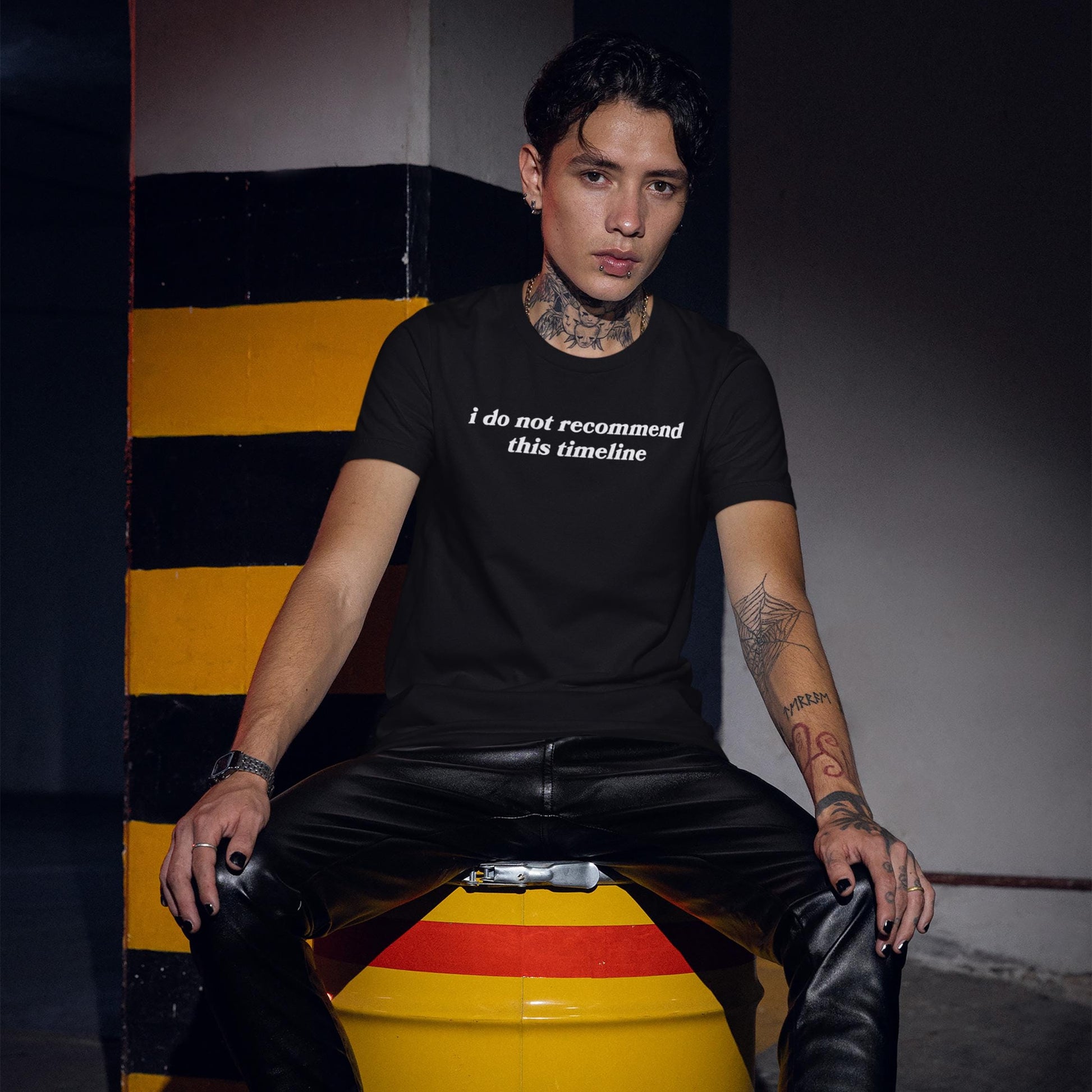 Model wearing black Timeline T-shirt with white text "i do not recommend this timeline" sitting on a yellow and red barrel in dim lighting.