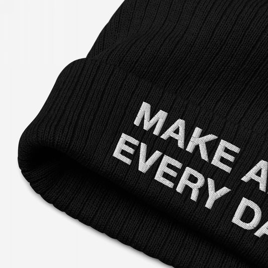 Make Art Every Day Beanie: Close-up of a black ribbed knit beanie with a folded cuff, featuring the embroidered words “MAKE A” and “EVERY D” in white block letters on the cuff.