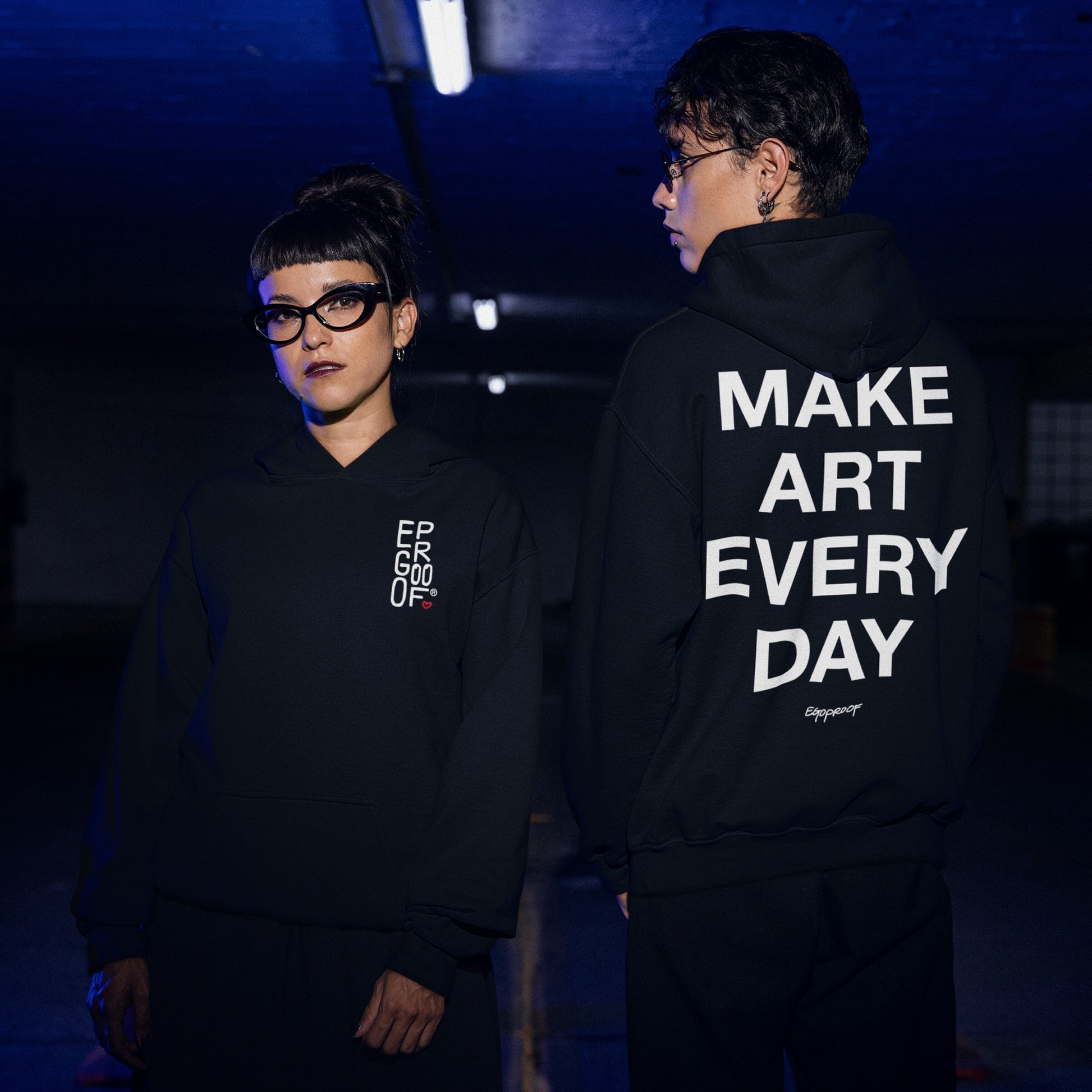 Two people in a dim garage wear the Make Art Every Day Hoodie; front features a subtle logo, back boldly states MAKE ART EVERY DAY, embodying a commitment to creativity.