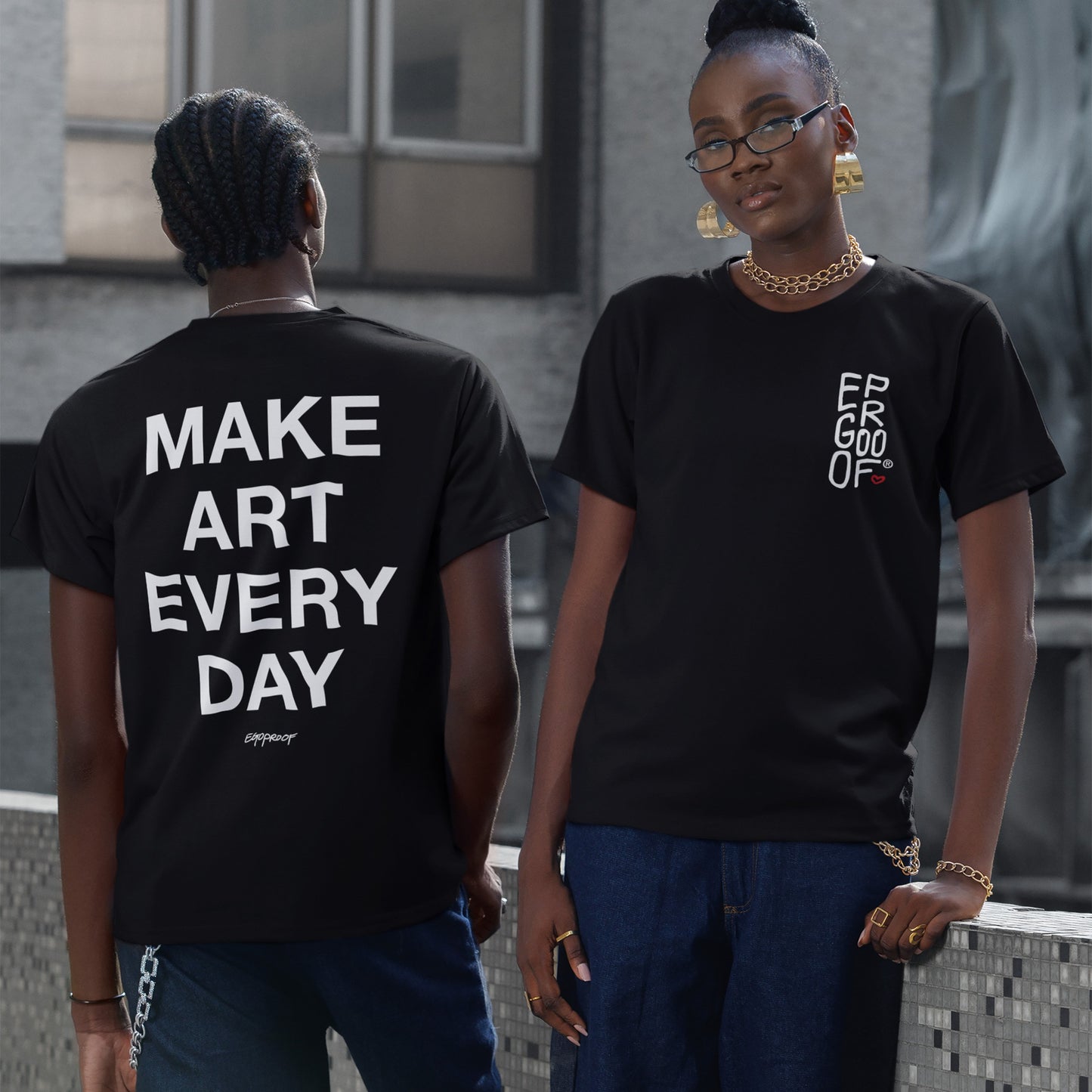 Models wearing the Make Art Every Day T-shirt EU show its bold slogan on the back and a stylized logo on the front, styled with denim and gold accessories in an urban setting.
