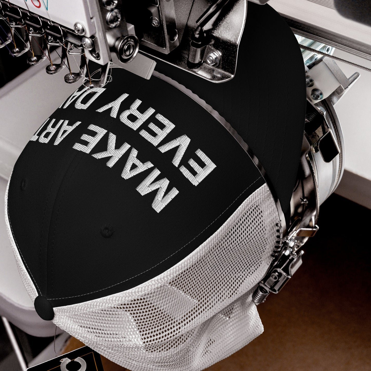 Make Art Every Day Hat being embroidered on an industrial machine, showcasing bold white lettering on the front panel of a black-and-white mesh trucker cap.