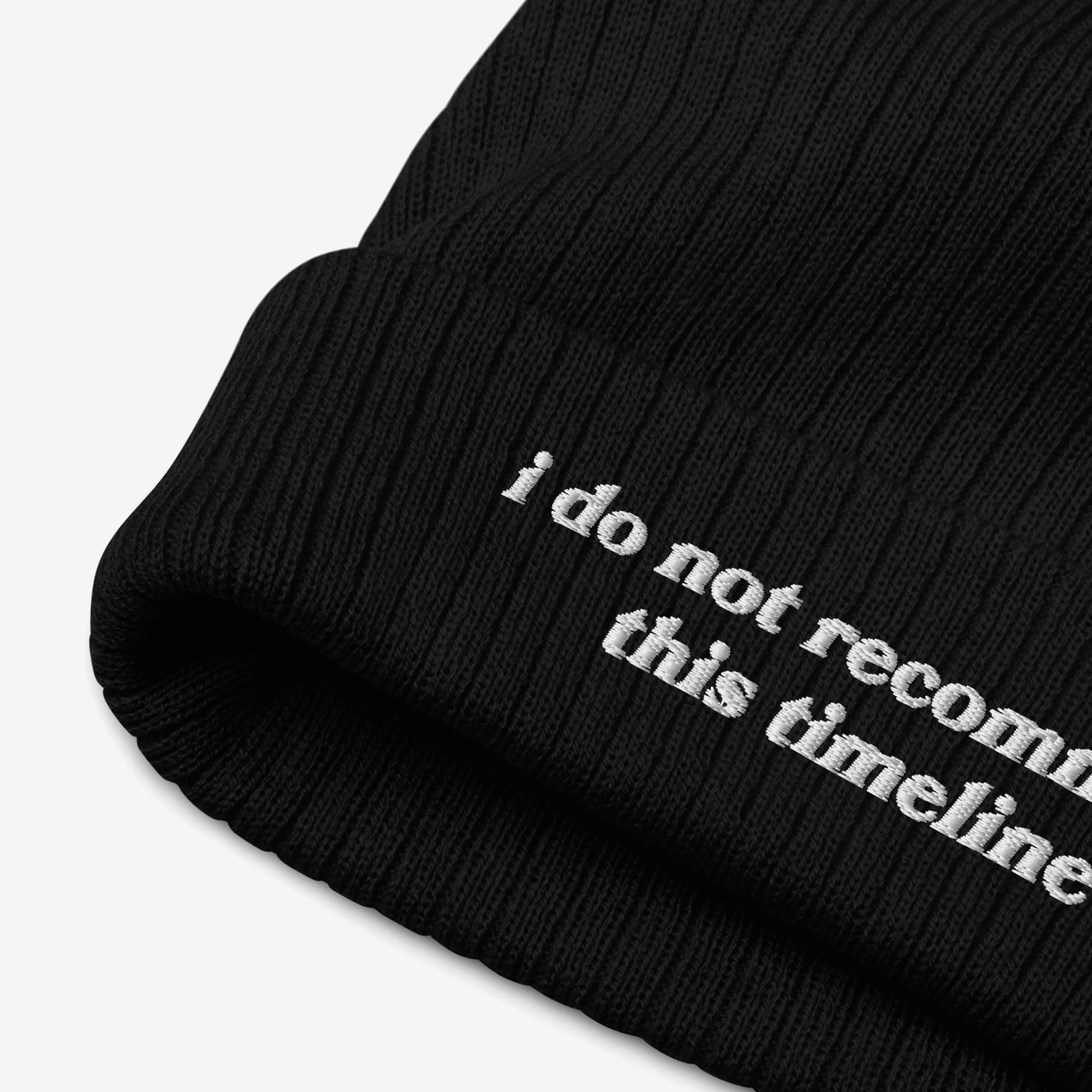 Close-up of the Timeline Hat, a black ribbed knit beanie with a folded cuff, featuring partial white embroidery: “i do not recommend this timeline.” Ideal for sarcastic flair amidst modern chaos.