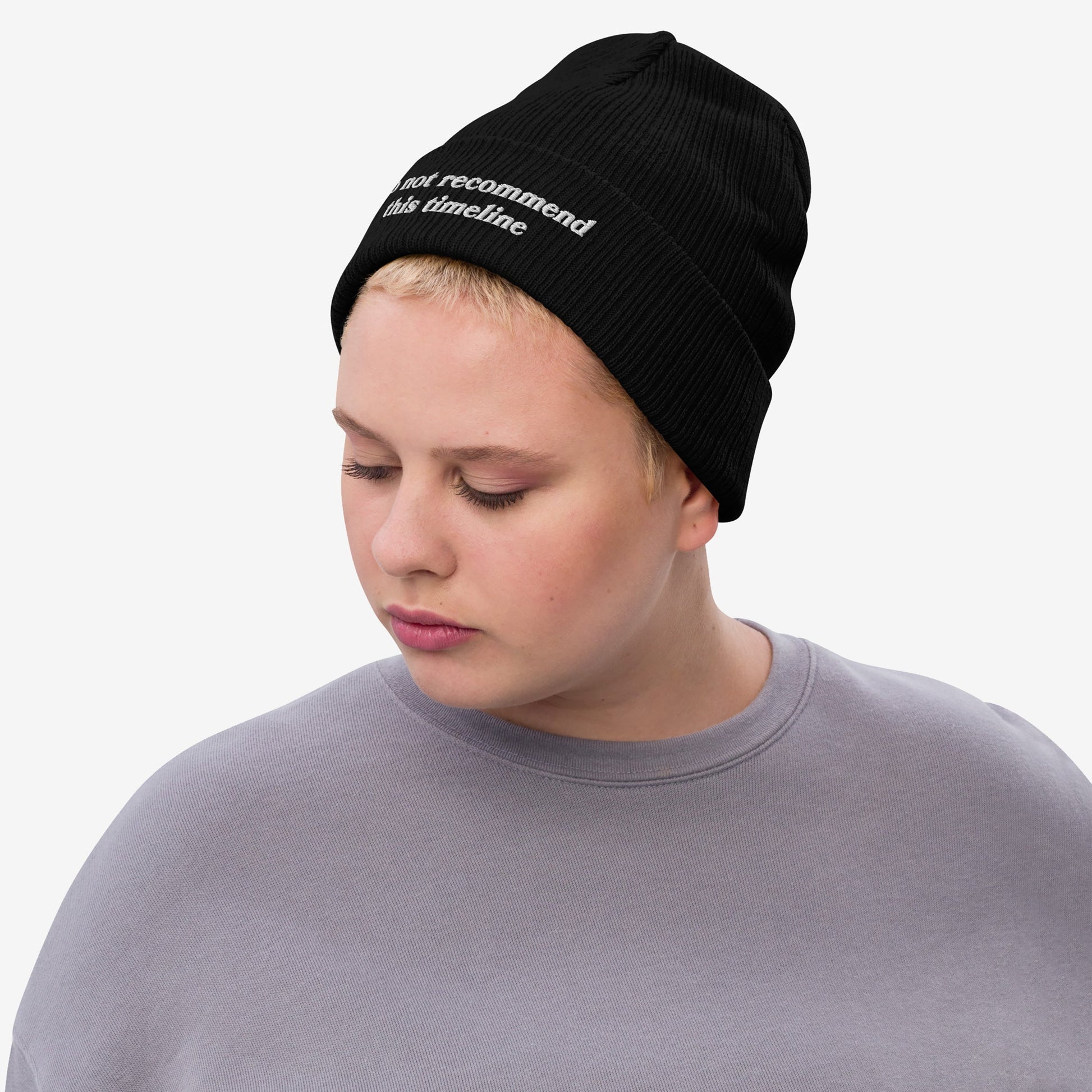 Person with short blond hair wears a light gray sweatshirt and a black Timeline Hat beanie, humorously embroidered with “do not recommend this timeline,” embodying Egoproof's statement-making, mental health-focused apparel.
