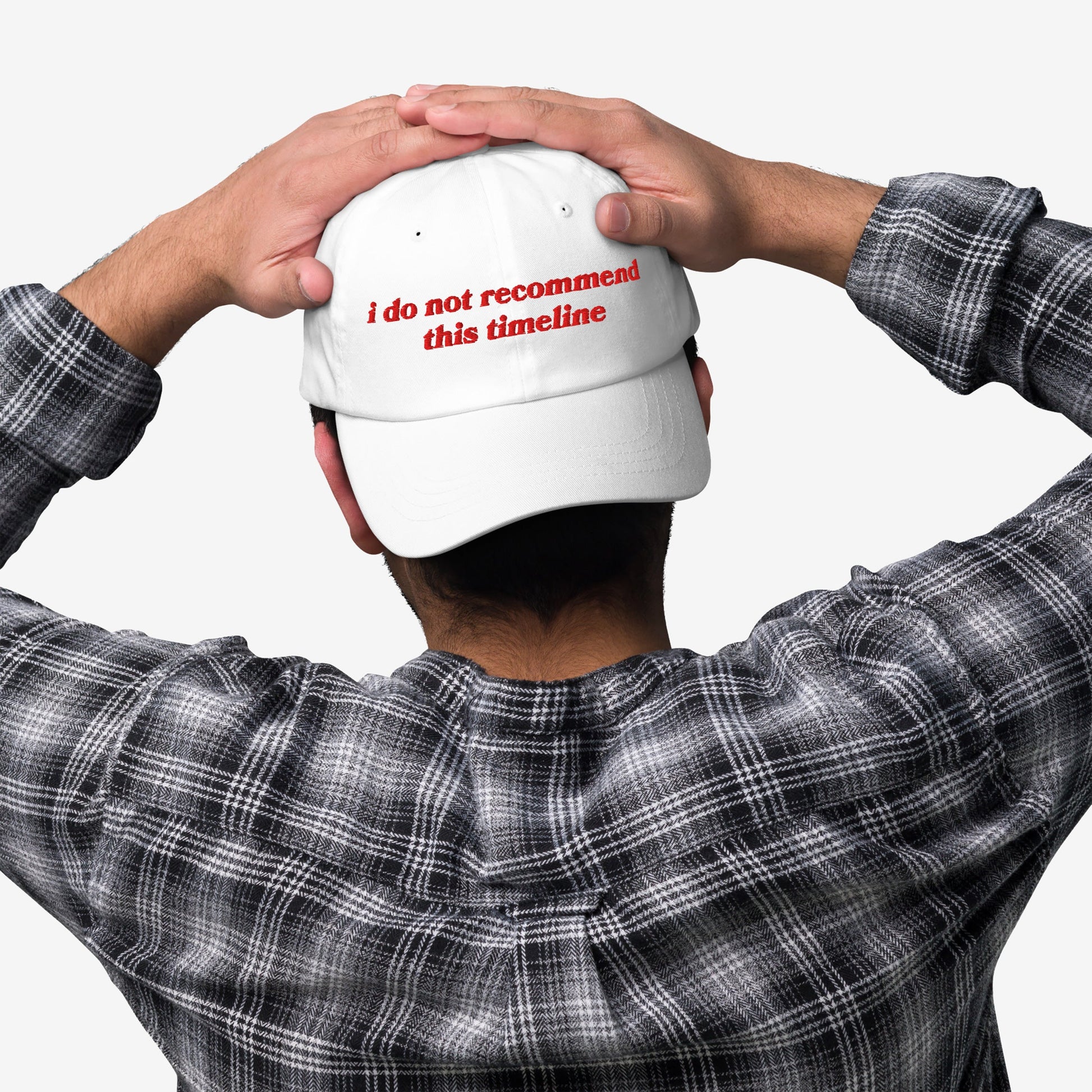 Person in a black-and-white plaid shirt and white cap with I Do Not Recommend This Timeline embroidered, hands on cap, showcasing Egoproof's statement piece against modern chaos.