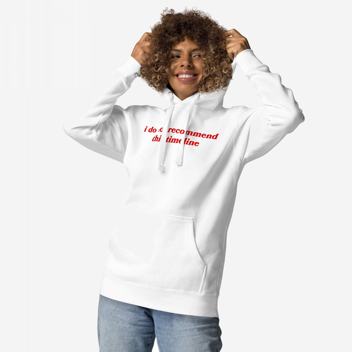 Person with short curly hair smiles while adjusting the hood of the Timeline Hoodie EU, featuring I do not recommend this timeline in red text, paired with light blue jeans.
