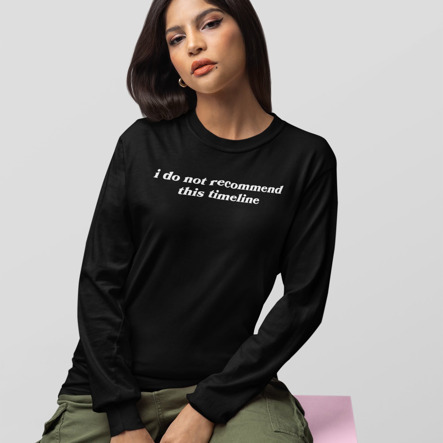 Woman wearing black Timeline Long Sleeve T-shirt with white "i do not recommend this timeline" text sitting against gray background.