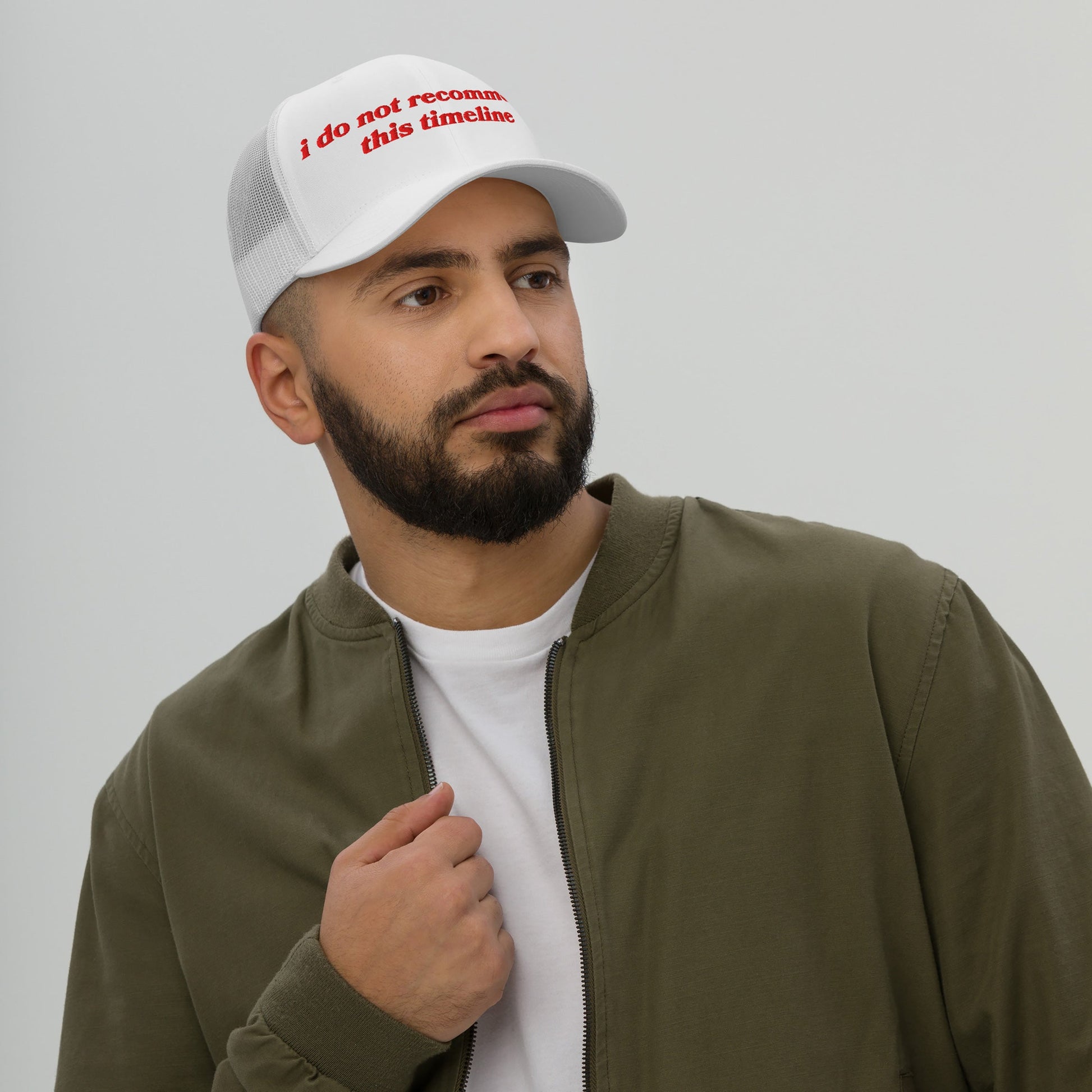 Man wearing the Timeline Hat, a white mesh-back trucker cap with red text i do not recommend this timeline, styled with a white T-shirt and olive-green jacket, embodying modern chaos and irony.