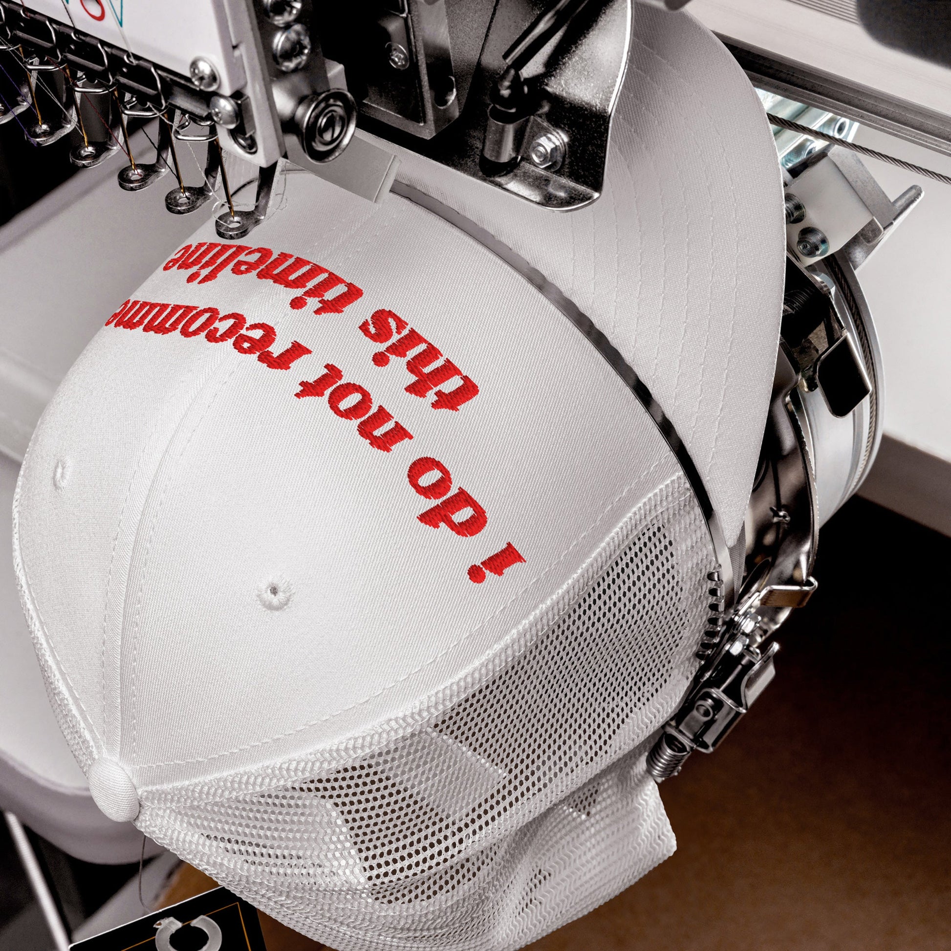 White mesh-back trucker cap, Timeline Hat, in an embroidery machine, red text being stitched on the front. A statement piece for those navigating modern chaos with irony, supporting mental health awareness.