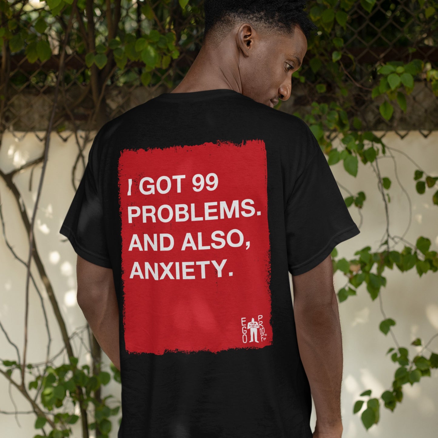 Person wearing the 99 Problems T-shirt, featuring a red rectangle with white text: I GOT 99 PROBLEMS. AND ALSO, ANXIETY. on the back. A small white EGOPROOF logo appears in the corner.