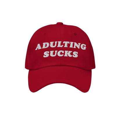 Red baseball-style cap with ADULTING SUCKS embroidered in white on front, representing Egoproof's relatable, bold apparel. Id