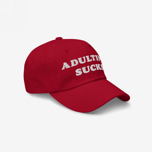 Red baseball-style cap with white embroidered lettering reading “ADULTING SUCKS,” the Adulting Sucks Hat. Ideal for expressing mental health struggles with style and honesty, supporting NAMI.