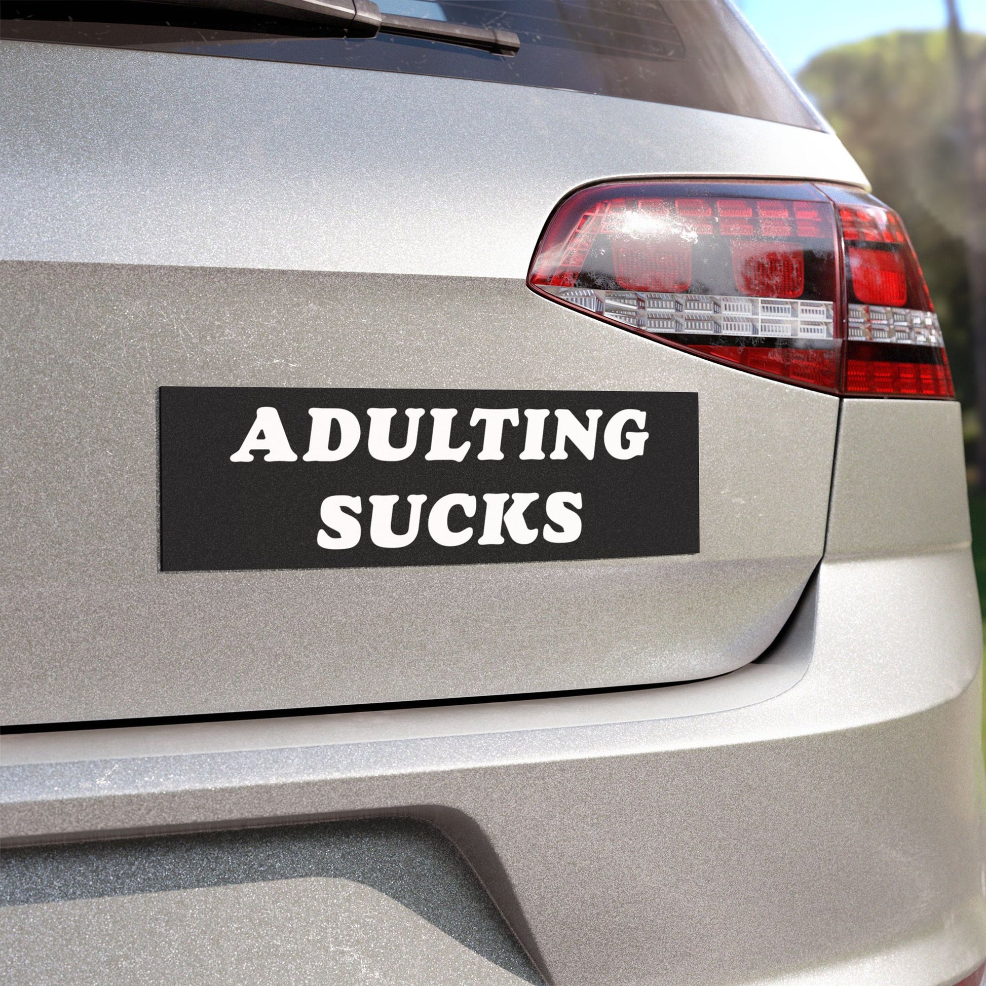 The rear of a silver hatchback displays the Adulting Sucks Sticker & Magnet with its bold slogan, encapsulating the everyday exhaustion of adulthood in a durable, matte black bumper sticker.