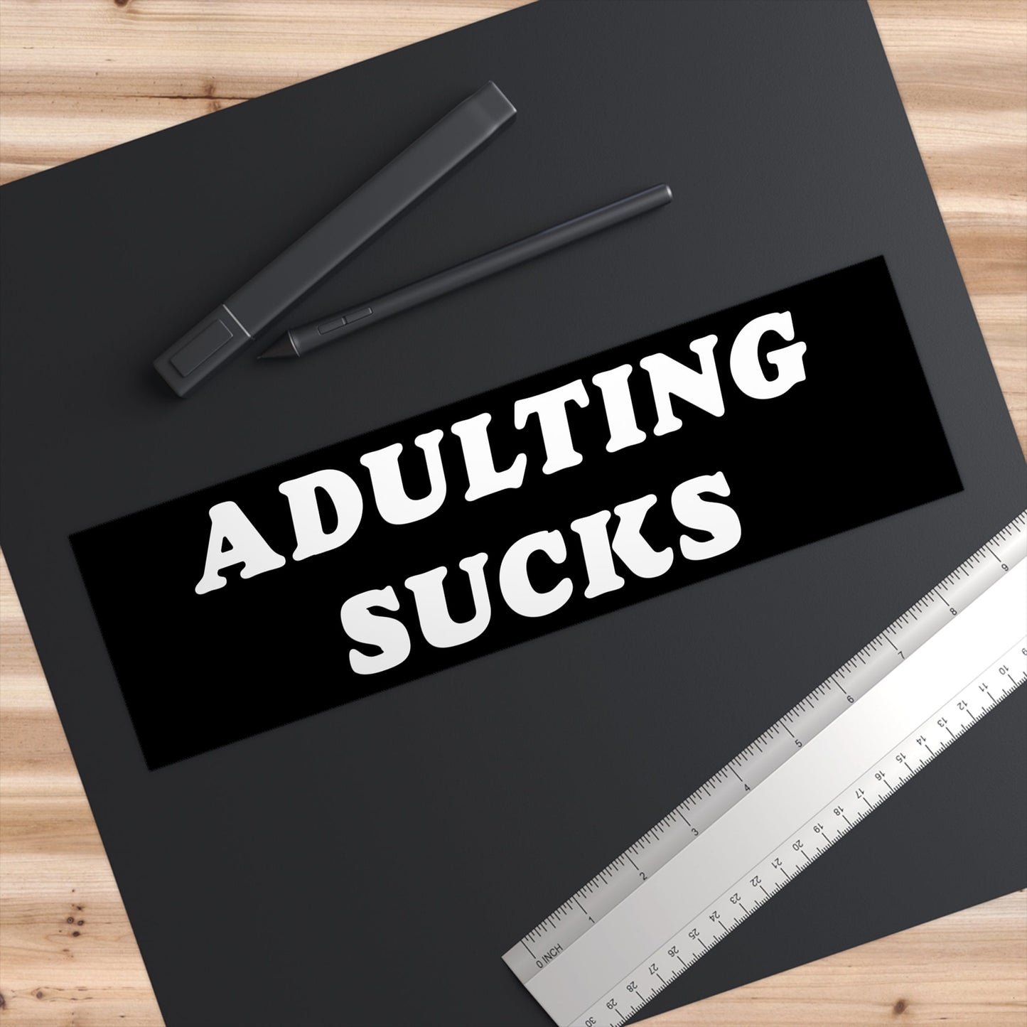 Adulting Sucks Sticker & Magnet displayed on a black desk mat with a digital pen and stainless-steel ruler, conveying a humorous take on adulthood's challenges. Perfect for cars, laptops, or workspace decor.