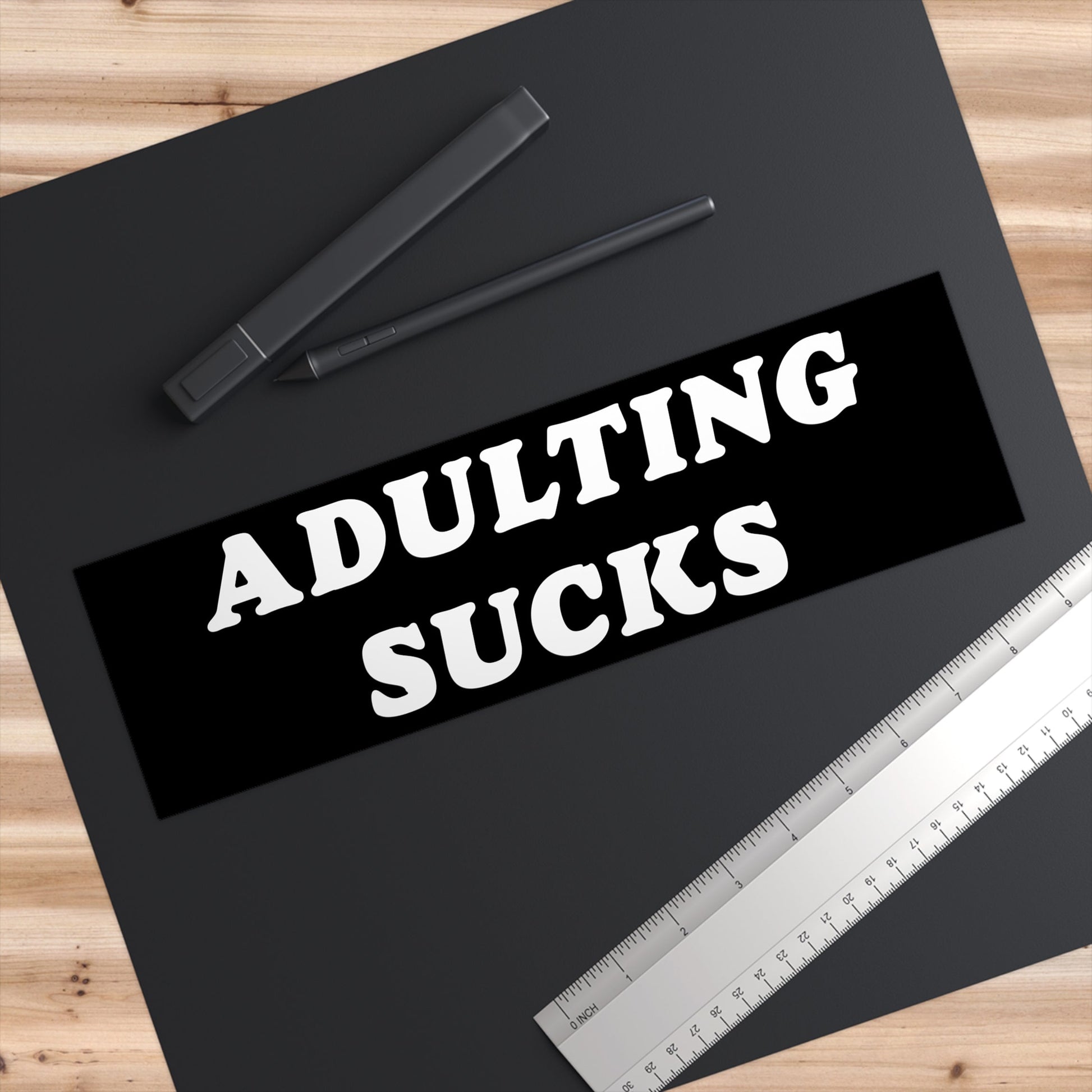 Adulting Sucks Sticker & Magnet displayed on a black desk mat with a digital pen and stainless-steel ruler, conveying a humorous take on adulthood's challenges. Perfect for cars, laptops, or workspace decor.