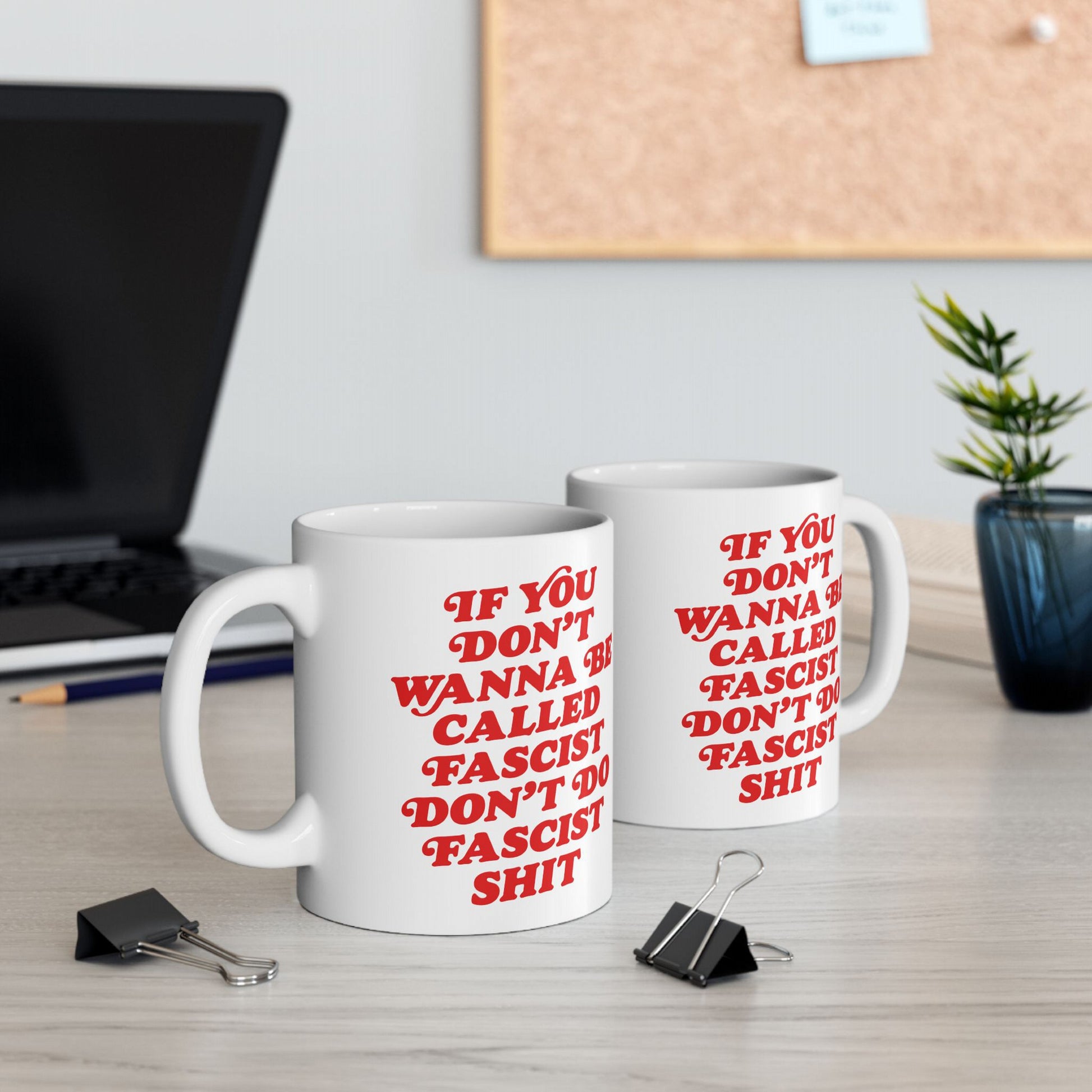 Anti-Fascist Mug with bold red slogan sits on a desk, surrounded by a laptop, pencil, binder clips, and a potted plant, embodying defiance and resistance in everyday life.
