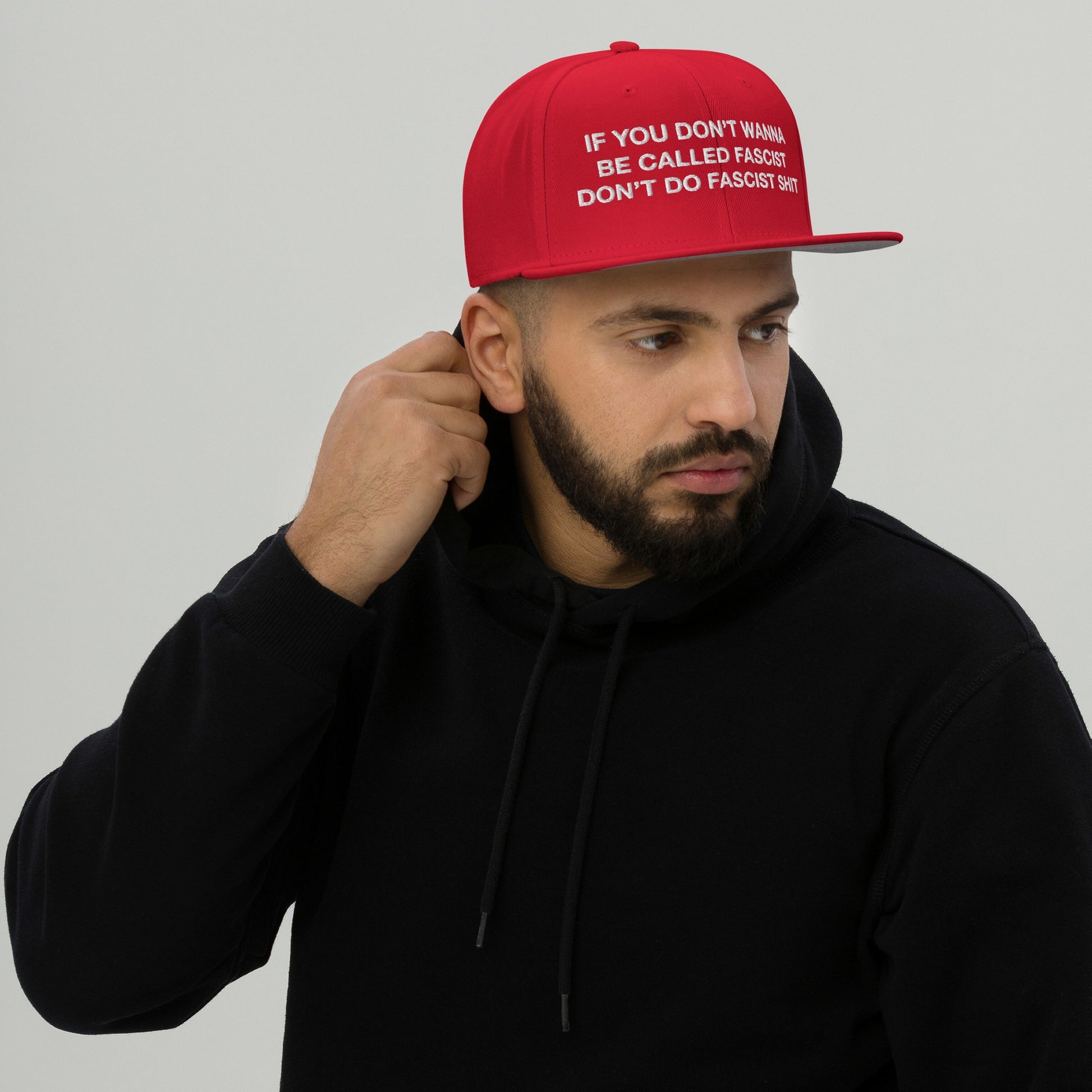 Young man in a black hoodie adjusts the Anti-Fascist Hat, a bold red snap-back cap with a powerful embroidered slogan promoting resistance against fascism.