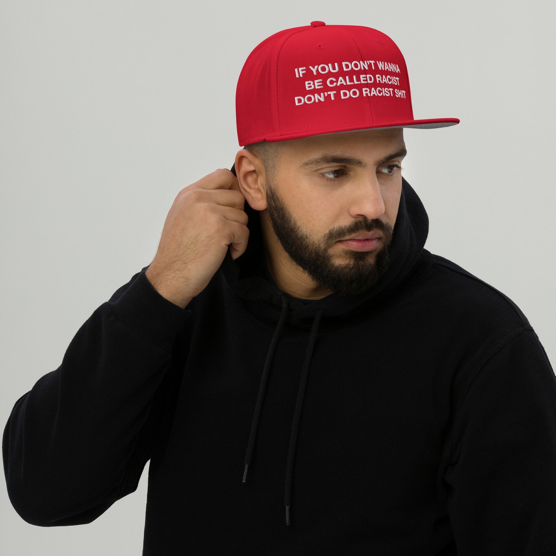 Anti-Racist Hat EU: A bold red cap with the slogan “IF YOU DON’T WANNA BE CALLED RACIST DON’T DO RACIST SHIT,” worn by a bearded man in a black hoodie.