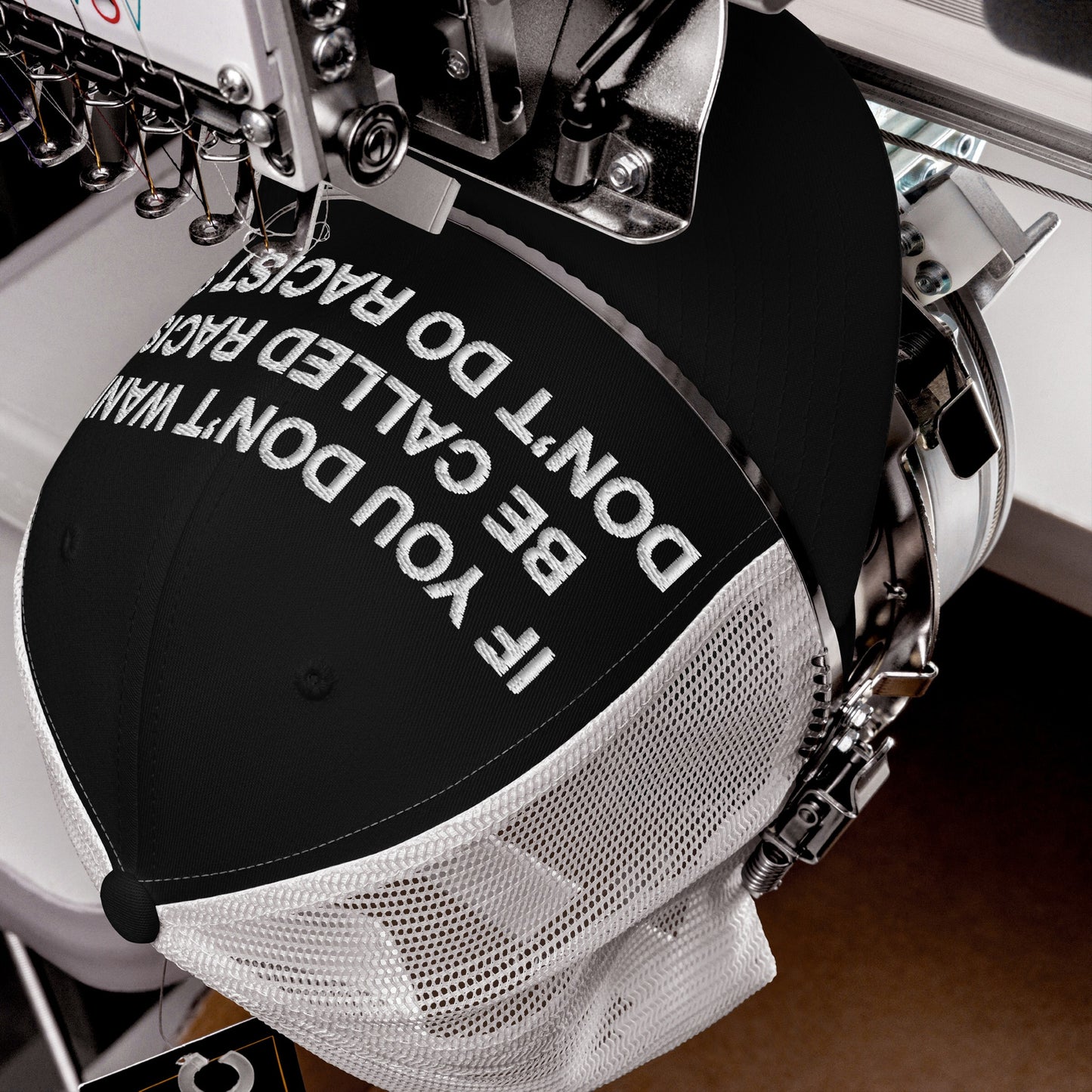Black-front, white-mesh back trucker cap titled Anti-Racist Hat EU is in an embroidery machine, stitching IF YOU DON'T WANT TO BE CALLED RACIST, DON'T DO RACISM, reflecting a bold anti-racist statement.