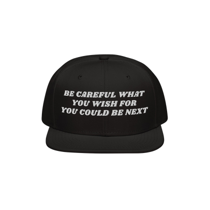 Black flat-brim snapback cap with bold white embroidery stating, Be Careful What You Wish For, You Could Be Next. The Be Care