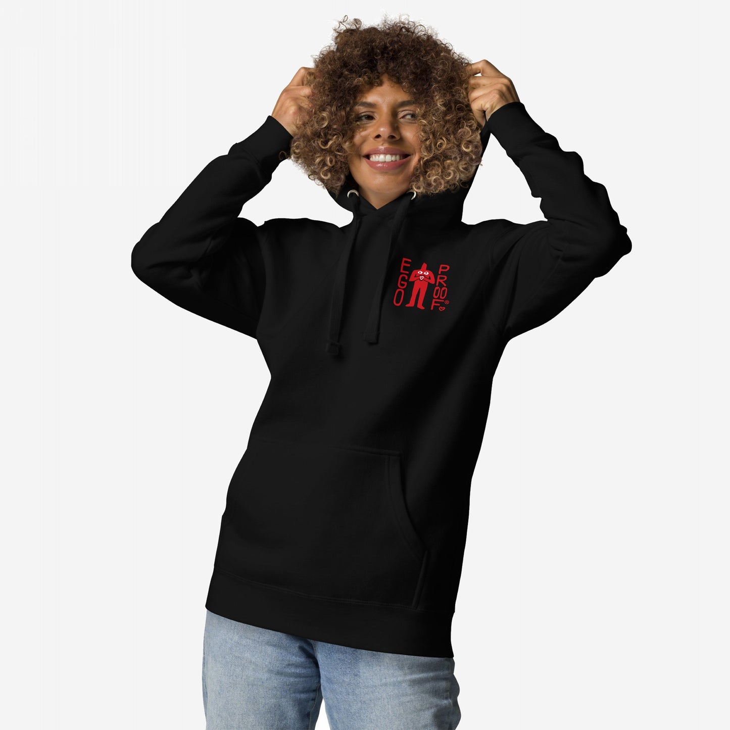 Curly-haired person wears the Fucks To Give Hoodie EU, featuring a small red “EGO PROOF” graphic on front and bold statement on back, holding drawstrings while smiling in light-wash jeans.